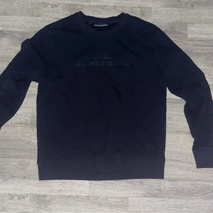 Mörkblå sweatshirt från J.Lindeberg - En stilren mörkblå sweatshirt från J.Lindeberg med diskret logga framtill. Tröjan har rund halsringning och långa ärmar. Perfekt för en avslappnad och snygg look.