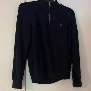 Svart stickad tröja med dragkedja från Gant - Svart stickad tröja från Gant med halv dragkedja framtill och diskret logga på bröstet. 
