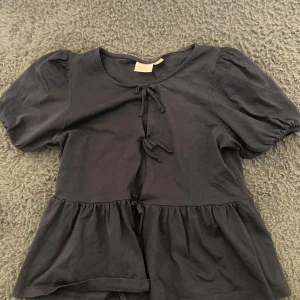 Svart blus med knyt och puffärm - Säljer en svart blus med korta puffärmar och knyt framtill. Blusen har en lätt volang nedtill och är tillverkad i mjukt bomullstyg. Perfekt för dig som gillar feminina detaljer och enkel stil. Aldrig använd!