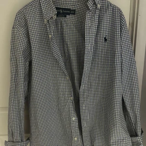Rutig skjorta från Ralph Lauren - Klassisk rutig skjorta från Ralph Lauren i blått och vitt. Skjortan har lång ärm, knappar framtill och den ikoniska broderade loggan på framsidan.
