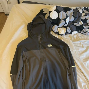 Svart och mörkgrå vindjacka från The North Face - Säljer en snygg svart och mörkgrå vindjacka från The North Face med huva och dragkedja. Jackan har en sportig look och är perfekt för utomhusaktiviteter. Logga på bröstet och reflekterande detalj på ärmen.
