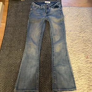 Blå bootcut jeans från Telepatía med detaljerad bakficka - Säljer ett par blå bootcut jeans från Telepatía med snygga slitningar och kontrastsömmar. En knapp är också borta:(. Bakfickan har broderade vingar och glittriga detaljer som ger extra attityd. Klassisk femficksmodell med låg midja och dragkedja. 