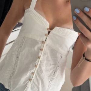 Vit linnetopp med knappar - Säljer en söt vit linnetopp med smala axelband och dekorativa knappar framtill. Toppen har fina spetsdetaljer och är perfekt för varma dagar. Lätt och luftig känsla. Nästan aldrig använd 