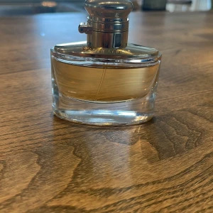 Parfym från Ralph Lauren med silverfärgat lock - Elegant parfymflaska från Ralph Lauren med ett stilrent, ovalt glas och ett blankt silverfärgat lock. Flaskan har en gyllene vätska inuti och RL-loggan på locket. Perfekt för dig som gillar klassisk och lyxig design.