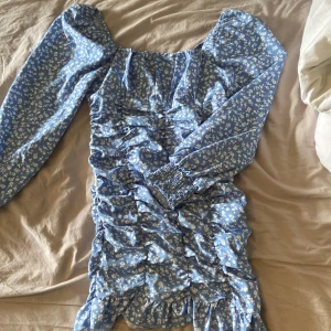 Blå blommig klänning med volangdetaljer - Supersöt blå klänning med vita småblommor och volangdetaljer. Klänningen har långa puffärmar med resår vid handlederna och smockad effekt längs hela framsidan. Perfekt för vår och sommar!