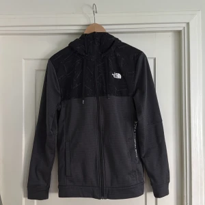 Svart hoodiejacka från The North Face - Säljer en svart hoodiejacka från The North Face med dragkedja och justerbar huva. Jackan har diskret mönster på axelpartiet och två fickor framtill. Perfekt för lager-på-lager och vardagsstil.