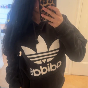 Svart hoodie från Adidas med stor logga - Svart hoodie från Adidas med stor vit trefoil-logga och text på bröstet. 