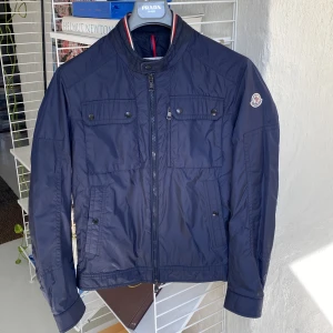 Mörkblå vindjacka från Moncler - Snygg mörkblå vindjacka från Moncler med ståkrage och dragkedja framtill. Jackan har två bröstfickor med knappar och Moncler-logga på ärmen. Perfekt för vår och sommar. Väldigt sparsamt använd. Superbra skick. Storlek 3. 