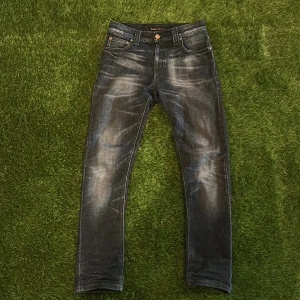 Mörk blåa jeans från Nudie Jeans - Säljer ett par mörk blåa jeans från Nudie Jeans med klassisk femficksdesign och snygga slitningar framtill och baktill. Jeansen har raka ben och normal passform, samt detaljer med nitar och knappstängning.