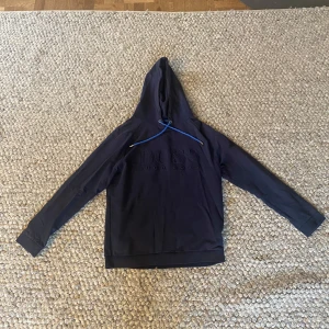 Mörkblå hoodie från BOSS - Säljer en mörkblå hoodie från BOSS med broderad logga på bröstet och blå dragsko i huvan. Tröjan har lång ärm och klassisk passform. Perfekt för en stilren och avslappnad look.
