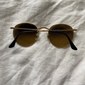 Runda solglasögon från Ray-Ban - Snygga runda solglasögon från Ray-Ban med guldfärgad metallbåge och bruna polariserade glas. Tunn design med svarta skalmar och klassisk Ray-Ban-logga på linsen. Perfekt accessoar för soliga dagar.