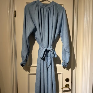 Ljusblå långklänning med bälte - Säljer en ljusblå långklänning med lång ärm och rund hals. Klänningen har ett avtagbart bälte i midjan och resår vid ärmsluten. Materialet känns som bomull eller linne och den har en enkel, stilren look.