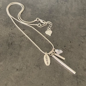 Silverfärgat halsband med berlocker och texten GEMINI - Säljer ett silverfärgat halsband med flera berlocker: en oval med texten GEMINI, två stavar och en diamant. Kedjan har även ett litet hjärta vid låset med samma text. Perfekt för dig som är tvilling i stjärntecken.
