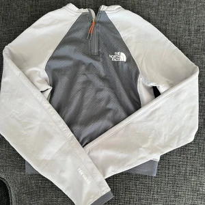 Grå långärmad tröja från The North Face - Säljer en grå långärmad tröja till träning eller vardag från The North Face med halv dragkedja och orange detalj. Tröjan har mjukt fleecefoder på insidan men är tunn och lätt. Oanvänd.