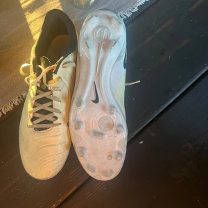 Nike fotbollsskor  - Säljer ett par vita Nike fotbollsskor med svarta detaljer och snörning. Skorna har rund tå och platt sula med dobbar, perfekta för spel på gräsplan. Klassisk design med sportig känsla.