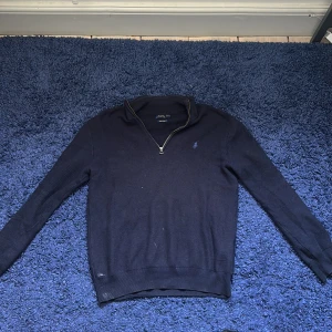 Mörkblå stickad tröja med dragkedja från Polo Ralph Lauren - Säljer en mörkblå stickad tröja från Polo Ralph Lauren med halv dragkedja och broderad logga på bröstet. Tröjan är synlig använd och jag personligen gillar inte passformen, råkade spilla lite färg på tröjan som inte går bort i tvätten därför billigt pris, fraktas snabbt och pris kan diskuteras vid snabbt köp.
