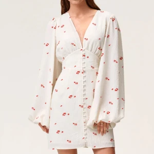 Vit klänning med 🍒 och knappar - Klänningen är oanvänd, prislappen är kvar! Säljer då jag glömde skicka tillbaka. Superfin vit kortklänning med små röda blommor, v-ringning och långa puffärmar. Klänningen har knappar framtill och markerad midja för en snygg siluett. Perfekt för sommarens alla tillfällen!