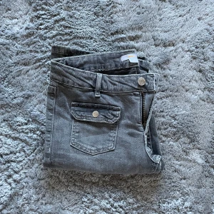 Grå bootcut jeans med fickdetaljer - Säljer ett par grå jeans med bootcut passform och snygga fickor framtill med lock och knapp. Jeansen har dragkedja och bälteshällor. Perfekta för dig som gillar klassisk stil med en twist.