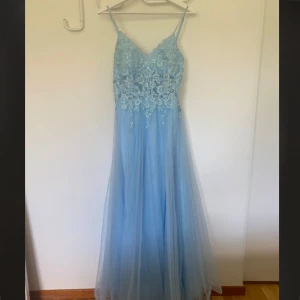 Ljusblå långklänning med spets och tyll - Säljer en ljusblå långklänning med smala axelband och v-ringning. Överdelen har vacker spets och broderade detaljer, medan kjolen är i flera lager tyll för en drömmig look. Perfekt för bal eller festliga tillfällen. Nypris 2600kr