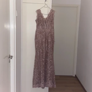 Rosa långklänning  - Elegant långklänning i ljusrosa spets med v-ringad hals och ärmlös design. Klänningen har fina broderade detaljer och en figurnära passform upptill som faller snyggt längs kroppen. Perfekt för bal eller festliga tillfällen.