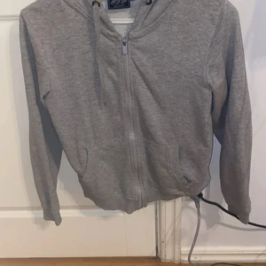 Grå hoodie med dragkedja - Grå hoodie med dragkedja framtill och huva med snören. Tröjan har långa ärmar och två fickor framtill. Perfekt för en avslappnad stil.