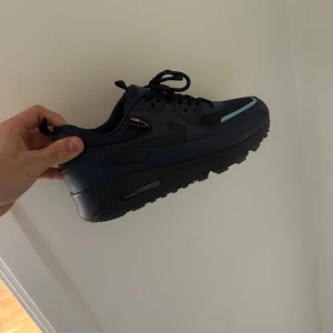 Nike Air Max 90 sneakers i mörkblått - Säljer ett par Nike Air Max 90 GORE-TEX sneakers i mörkblått med svarta detaljer och ljusblå accent på tån. Skorna har snörning och en robust, platt sula. Perfekta för dig som vill ha stilrena och vattentåliga sneakers.