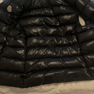 Svart dunväst från Moncler - Säljer en svart, glansig dunväst från Moncler med klassisk quiltad design. Västen har dragkedja framtill och Moncler-logga på bröstet. Perfekt för lager-på-lager och håller dig varm under kyliga dagar.