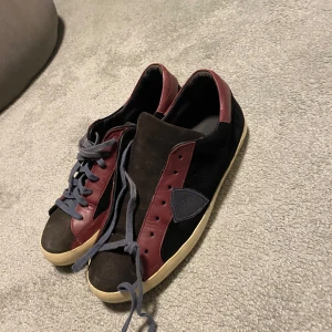 Philippe Model sneakers i svart och vinrött - Snygga sneakers från Philippe Model i svart mocka med vinröda detaljer och grå snörning. Skorna har en beige sula och en ikonisk sköld på sidan. Perfekta för dig som gillar stilrena och unika sneakers.