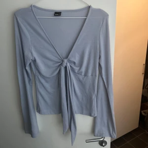 Ljusblå långärmad topp med knyt - Trendig topp från Gina med lång ärm och djup v-ringning. Toppen har en snygg knytning framtill. Använd 1 gång. 