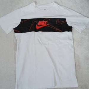 Vit t-shirt från Nike med svart och orange logga - Säljer en vit t-shirt från Nike med en svart panel över bröstet och röd Nike-logga. T-shirten har klassisk passform och är tillverkad i mjuk bomull. Perfekt för en sportig och avslappnad stil.