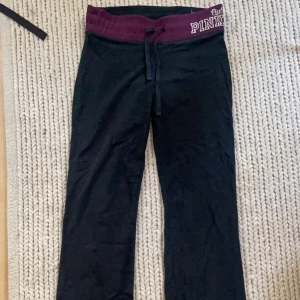 Svarta yogapants från PINK - Svarta yogapants från PINK med vinröd midja och snörning. Byxorna har en avslappnad passform och resår i midjan med tryckt logga. Perfekta för en chill dag hemma eller till träningen.
