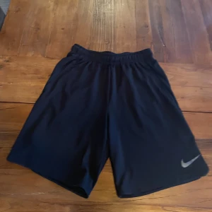 Svarta träningsshorts från Nike - Svarta träningsshorts från Nike med elastisk midja och snörning. Shortsen har en klassisk passform och är tillverkade i ett lätt och bekvämt polyestermaterial. Perfekta för gymmet eller löprundan. Nike-logga i silver på ena benet.