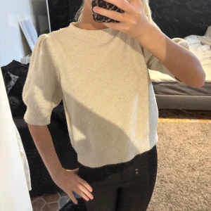Beige stickad tröja med puffärm - Säljer en beige stickad tröja med korta puffärmar och rund halsringning. Tröjan har ribbade muddar vid ärmslut och nederkant, vilket ger en snygg passform. 