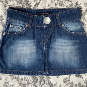 Jeanskjol från Six Jeans - Säljer en Vintage blå jeanskjol från Six Jeans med femficksmodell, stora bakfickor och silverfärgad lite större knapp framtilll. Kjolen är kort och passar från s-m skulle jag säga. Den är midrise,säljer pga att den är för stor😊