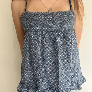 Blå mönstrad linnetopp med smock - Supersöt blå linnetopp med vitt småmönster och smock upptill. Toppen har tunna axelband som går att knyta och en lätt volangkant nertill. Perfekt för varma dagar!