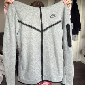 Grå zip-hoodie från Nike - Snygg grå zip-hoodie från Nike med svarta detaljer och dragkedja framtill. Jackan har huva, lång ärm och en praktisk ficka med dragkedja på ena ärmen. Perfekt för en sportig och avslappnad stil.