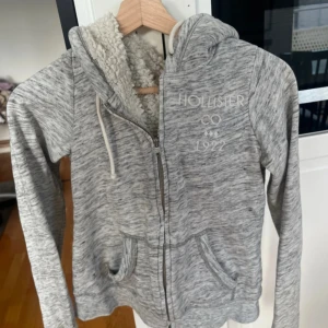 Grå zip-hoodie från Hollister med teddyfoder - Mysig gråmelerad hoodie från Hollister med dragkedja och vitt teddyfoder i luvan. Broderad logga på bröstet och två fickor framtill. Perfekt för kyliga dagar och enkel att matcha med det mesta.