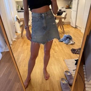 Blå jeanskjol från Zara Woman - Säljer en blå jeanskjol från Zara Woman med rå, fransig kant och slitna detaljer. Kjolen har klassisk femficksdesign, dragkedja och knapp framtill. Perfekt för en avslappnad och trendig look.