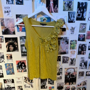 Grön ärmlös topp med blommor från Limited Collection - Säljer en grön ärmlös topp från Limited Collection med dekorativa rosetter på ena axeln. Toppen har en rund halsringning och är tillverkad i ett mjukt material. Perfekt för att liva upp garderoben med lite färg!