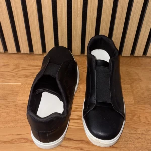 Svarta slip-on sneakers med vit sula - Svarta sneakers i syntetmaterial med elastiskt band över vristen istället för snörning. Skorna har en vit, platt sula och rund tå. Perfekta för en clean och enkel stil.
