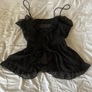 Svart volangtopp med knytband - Säljer en superfin svart volangtopp med tunna axelband och volangdetaljer. Toppen går att knyta på två ställen och är i ett genomskinligt material 