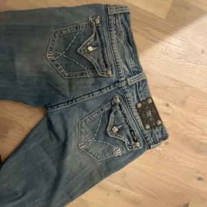 Miss Me jeans med slitningar och broderade bakfickor - Säljer ett par blå Miss Me jeans med snygga slitningar framtill och dekorativa broderade bakfickor med knappar. Jeansen har normal passform och klassisk femficksdesign. Perfekta för dig som gillar detaljer och en cool look.