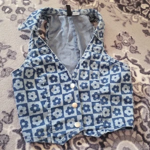 Blommig jeansväst från H&M - Säljer en blå jeansväst från H&M med blommigt mönster i rutigt. Västen har v-ringning, knappar framtill och en croppad passform. Perfekt för att ge din outfit en cool retro vibe🏵🌺