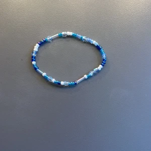 Blått och vitt pärlarmband - Säljer ett handgjort armband med små pärlor i olika nyanser av blått och vitt. Armbandet är elastiskt och består av både runda och avlånga pärlor som ger en fin variation. 