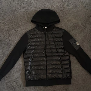 Svart pufferjacka med huva från Moncler - Snygg svart pufferjacka från Moncler med huva och dragkedja. Jackan har quiltad framsida, två fickor med dragkedja och en ärmficka med Moncler-logga. Perfekt för kyliga dagar och stilren look.med nfc tagg 