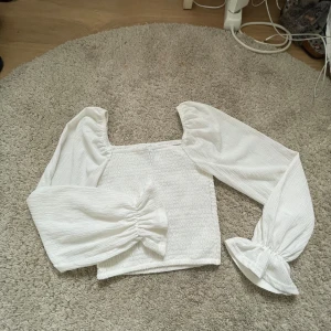 Vit blus med puffärmar från FB Sister - Säljer en söt vit blus från FB Sister i storlek XXS. Toppen har långa puffärmar med resår vid ärmslut och smockad kropp som ger en tight passform. Perfekt för dig som gillar romantisk och trendig stil!
