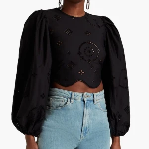 Svart croppad blus med broderade detaljer från Ganni - Trendig svart croppad blus från Ganni med broderade mönster och ballongärmar. Blusen har rund halsringning, dragkedja bak och vågig nederkant. Perfekt för att lyfta din outfit med unika detaljer.