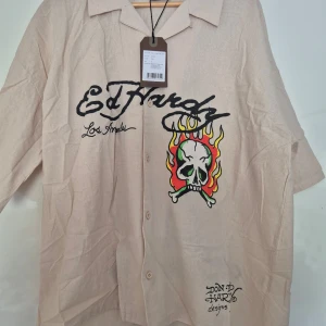 Beige skjorta med dödskalletryck från Ed Hardy - Säljer en beige Ed Hardy skjorta i relaxed fit med stora tryck av dödskalle och eld både fram och bak. Skjortan har korta ärmar, knappar framtill och klassisk krage. Perfekt för dig som gillar streetwear och unika motiv.