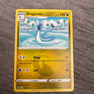 Dragonair Pokémonkort - Säljer ett Dragonair Pokémonkort från Sword & Shield-serien. Kortet har gul ram och visar Dragonair i en strandmiljö. Det är ett Stage 1-kort med 100 HP och har attackerna Wrap och Surf. Kortnummer 130/195.