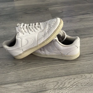 Vita Nike sneakers - Säljer ett par klassiska vita Nike sneakers med lågt skaft och snörning. Skorna har rund tå, perforerade detaljer framtill och platt sula. Perfekta för en clean och stilren look.